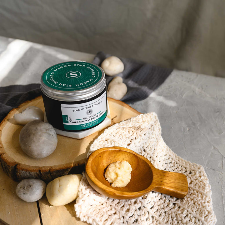 An 8 ounce container of Kate shea souffle sugar scrub is displayed with a wooden spoon showing the product, surrounded by natural elements like rocks and a knit washcloth, on a wooden surface.