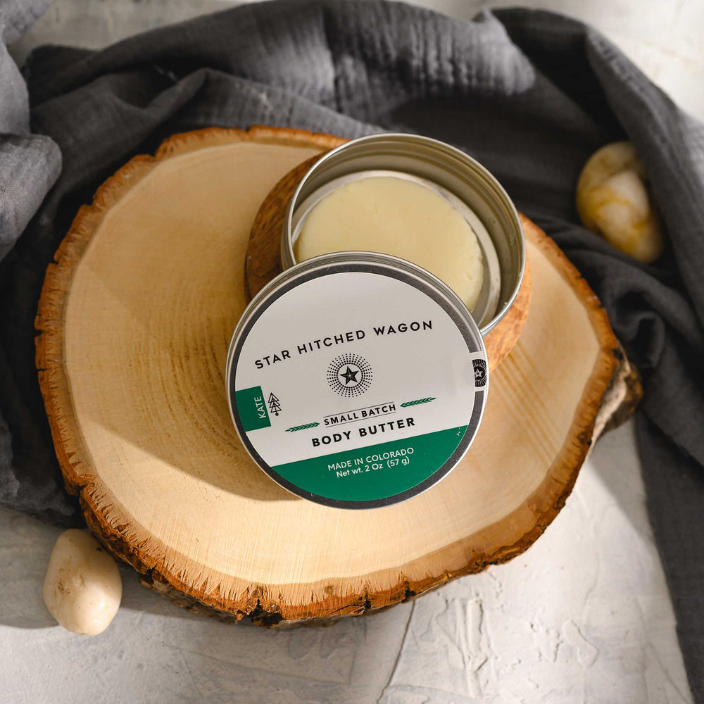 Kate body butter moisturizer in reusable tin, made with all natural oils and butters to soothe and protect dry skin. Forest-scented and handcrafted. Set on a wood and stone surface with pebbles and grey towel in background.