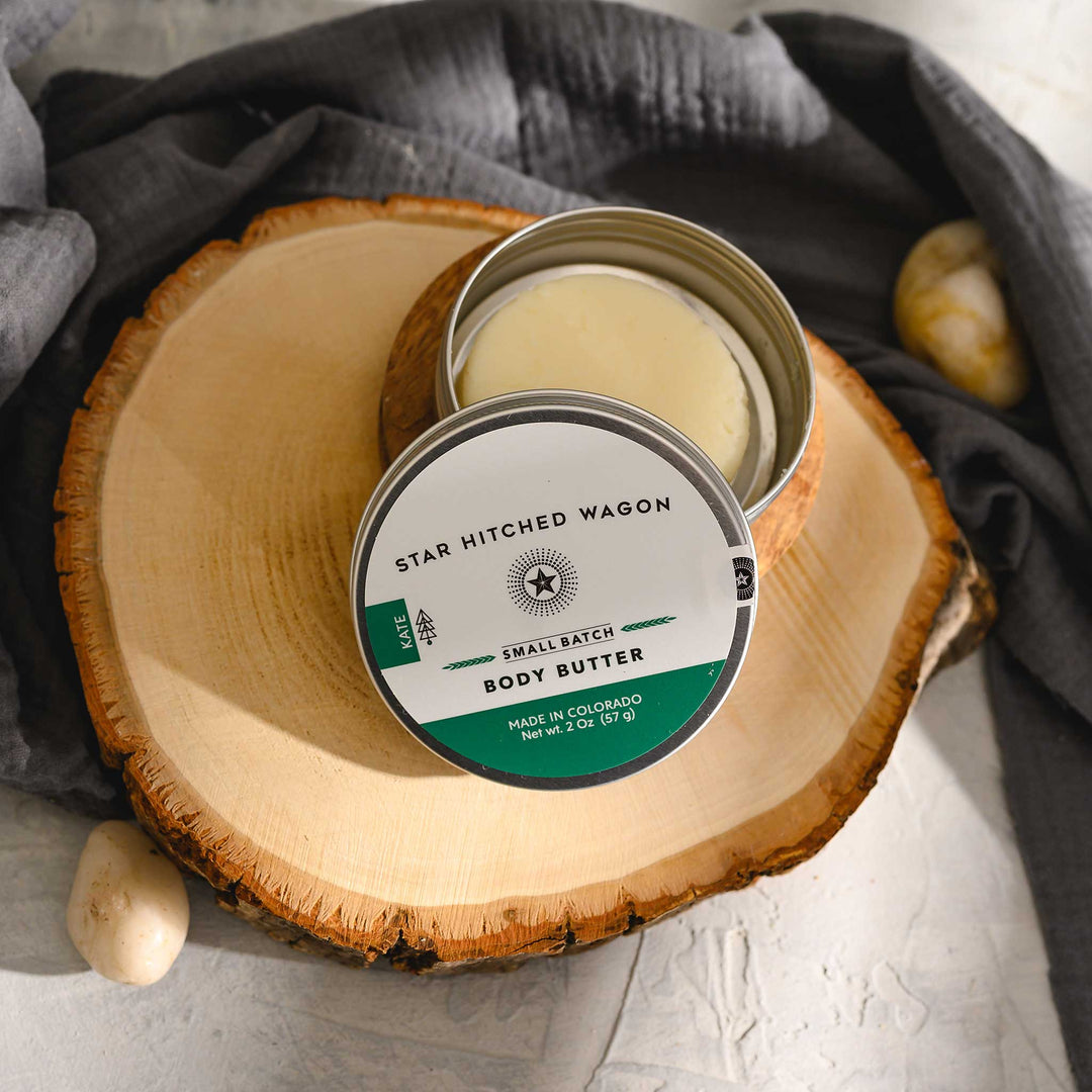 Kate body butter moisturizer in reusable tin, made with all natural oils and butters to soothe and protect dry skin. Forest-scented and handcrafted. Set on a wood and stone surface with pebbles and grey towel in background.