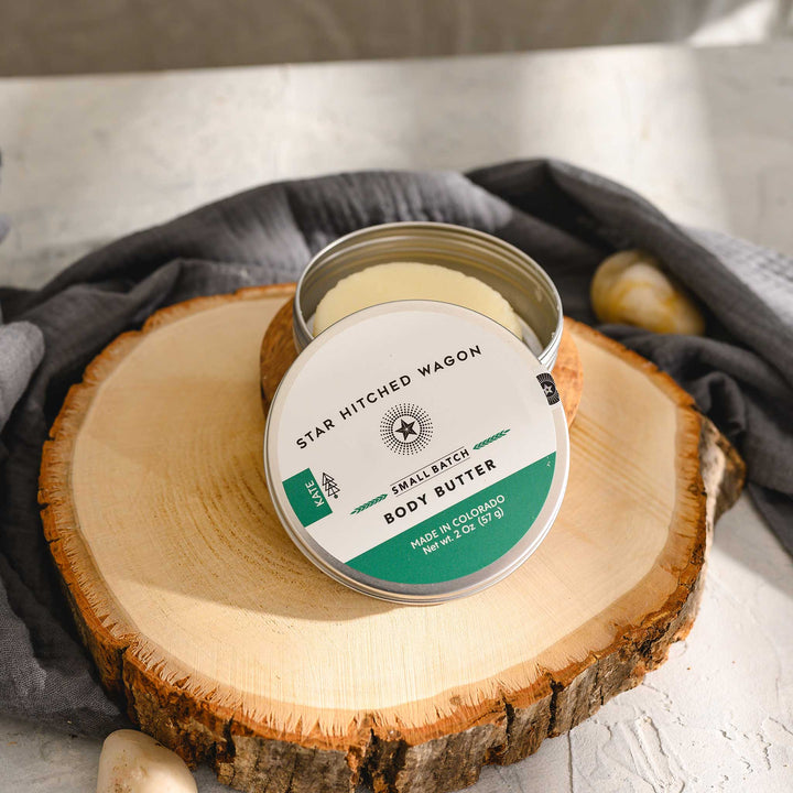 Kate body butter moisturizer in reusable tin, made with all natural oils and butters to soothe and protect dry skin. Forest-scented and handcrafted. Set on a wood and stone surface with pebbles and grey towel in background.