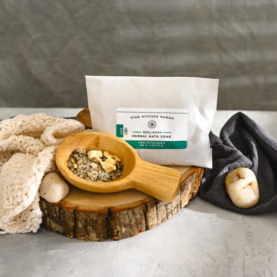 Wooden scoop filled with Kate Herbal Bath Soak next to full pouch on wooden slab next to a cream colored knit washcloth, a grey towel and pebbles.
