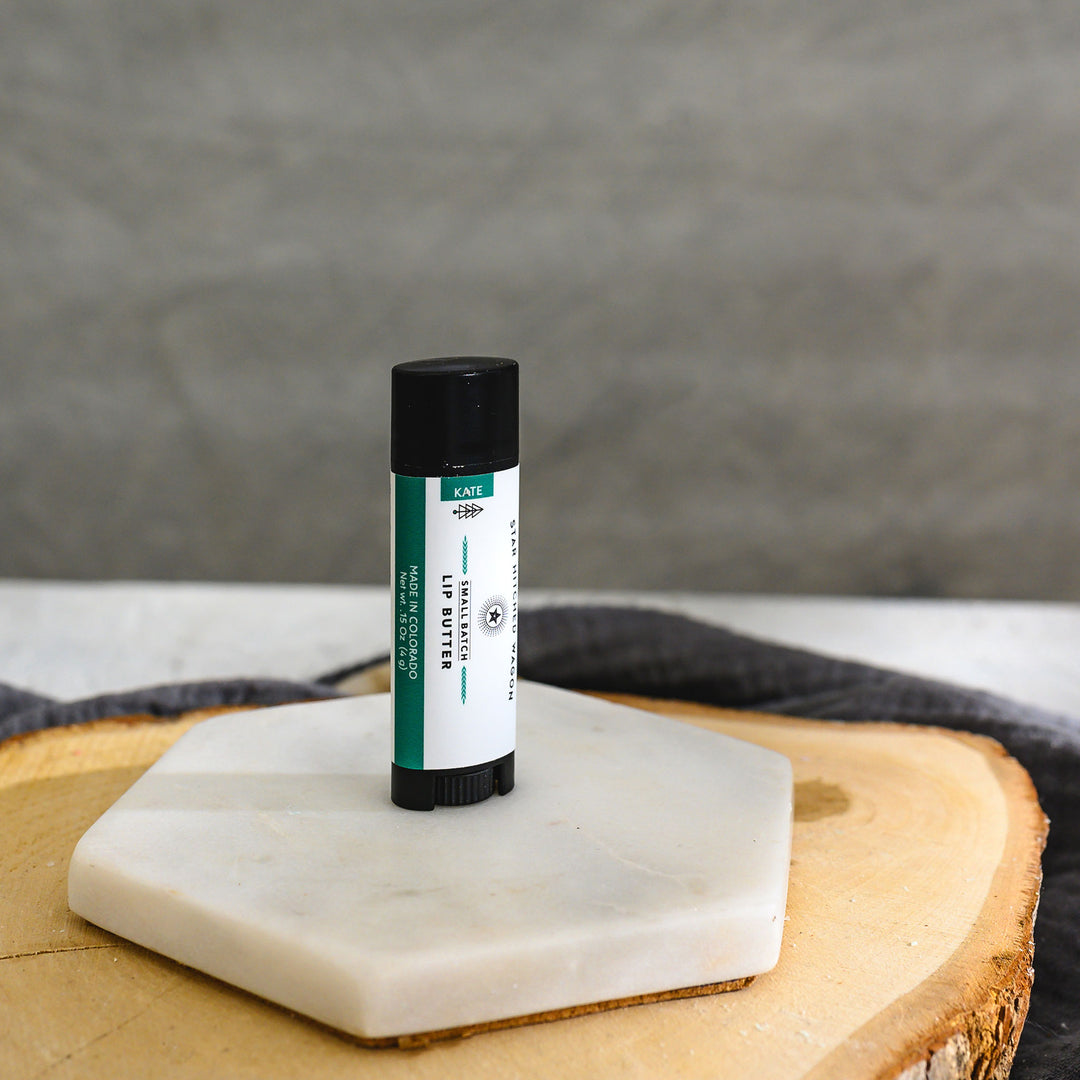 A tube of lip butter placed on a wood and stone surface with a grey towel n the background.