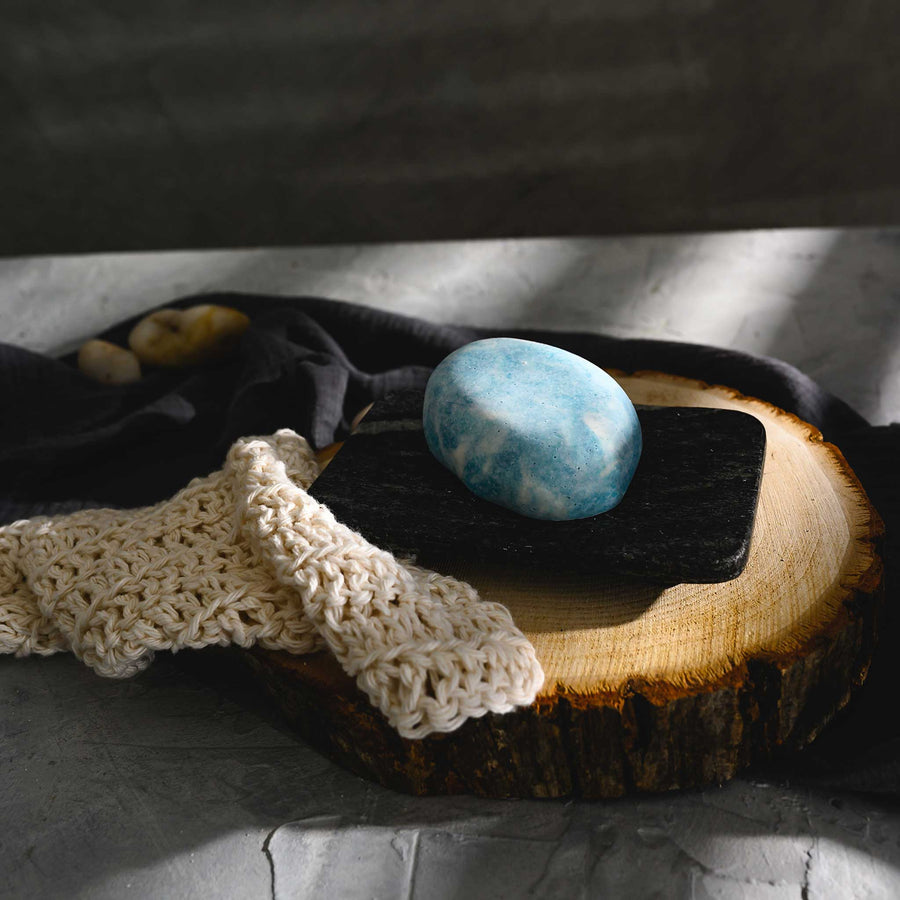 Kate river stone salt soap bar, placed on a wooden surface with a crocheted soap dish and a dark cloth in the background.
