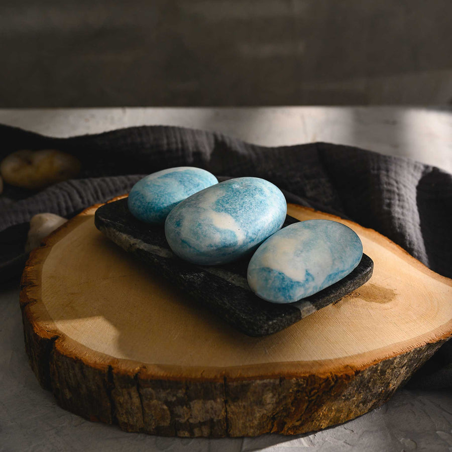 Kate river stone salt soap trio with a blue tint, placed on a rustic wooden dish, with a dark cloth in the background.