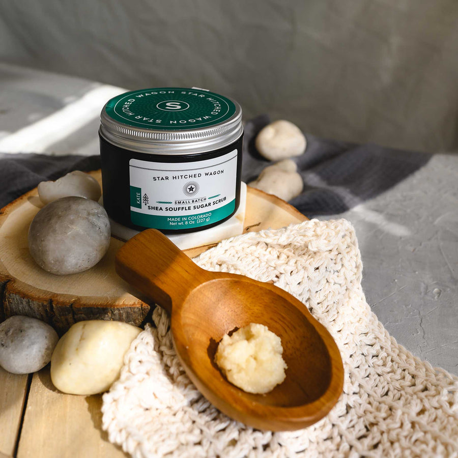 An 8 ounce container of Kate shea souffle sugar scrub is displayed with a wooden spoon showing the product, surrounded by natural elements like rocks and a knit washcloth, on a wooden surface.