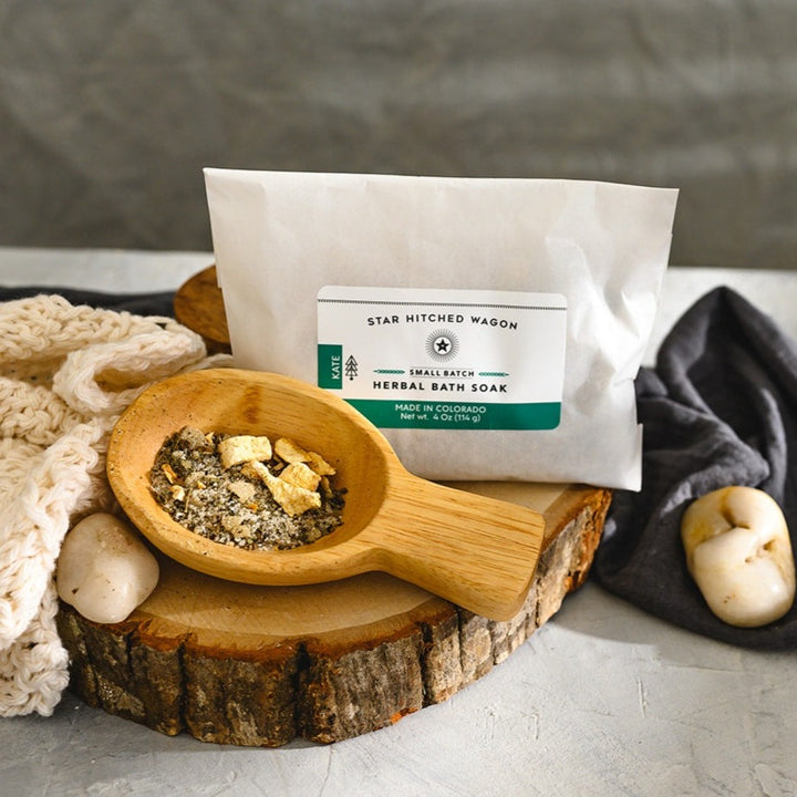Wooden scoop filled with Kate Herbal Bath Soak next to full pouch on wooden slab next to a cream colored knit washcloth, a grey towel and pebbles.