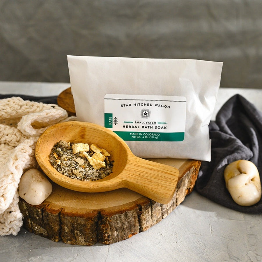Wooden scoop filled with Kate Herbal Bath Soak next to full pouch on wooden slab next to a cream colored knit washcloth, a grey towel and pebbles.