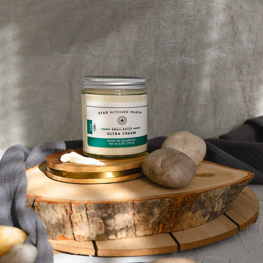 Kate Ultra Cream in a glass jar with an aluminum screw top lid, displayed on a wooden surface with a grey cloth and pebbles in the background.