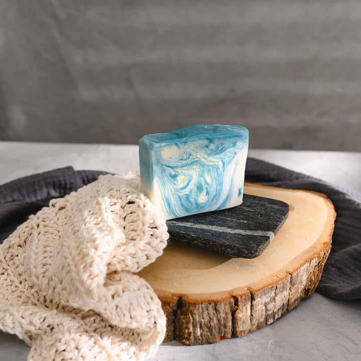 Kate essential oil soap bar with swirls of blue and white, placed on a stone and wood surface next to a cream colored knitted washcloth with a grey towel in the background.