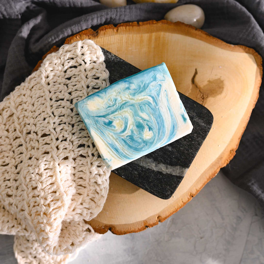 Kate essential oil soap bar with swirls of blue and white, placed on a wooden slice next to a cream colored knitted washcloth.