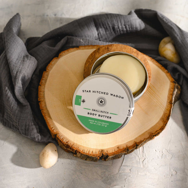 Liz body butter moisturizer in reusable tin, on stone and wood surface with pebbles and grey towel in background. Cool, minty, and refreshing.