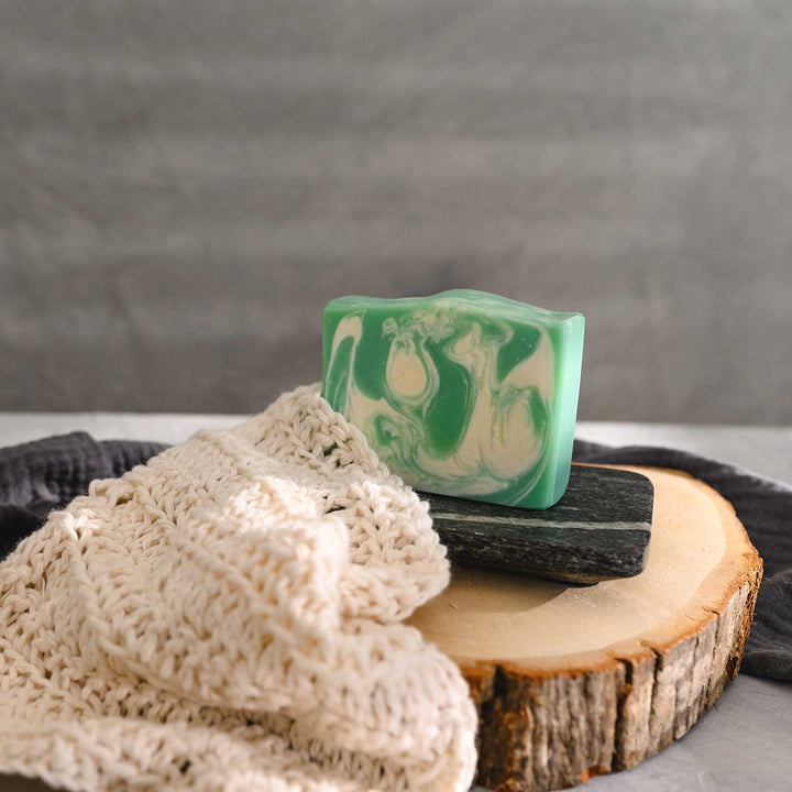 Liz essential oil soap bar with swirls of pale green and white, resting on a stone and wood surface with a cream colored washcloth beside it and a grey towel in the background.