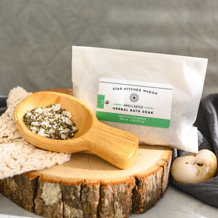 Wooden scoop filled with Liz Herbal Bath Soak next to full pouch on cream colored knit washcloth and wooden slab with grey towel and pebbles in background.
