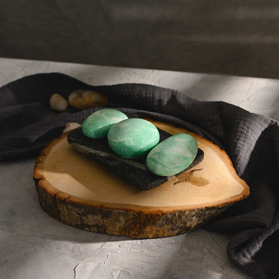 Liz river stone salt soap trio with a greenish tint, placed on a rustic wooden disc, with a dark gray cloth in the background.