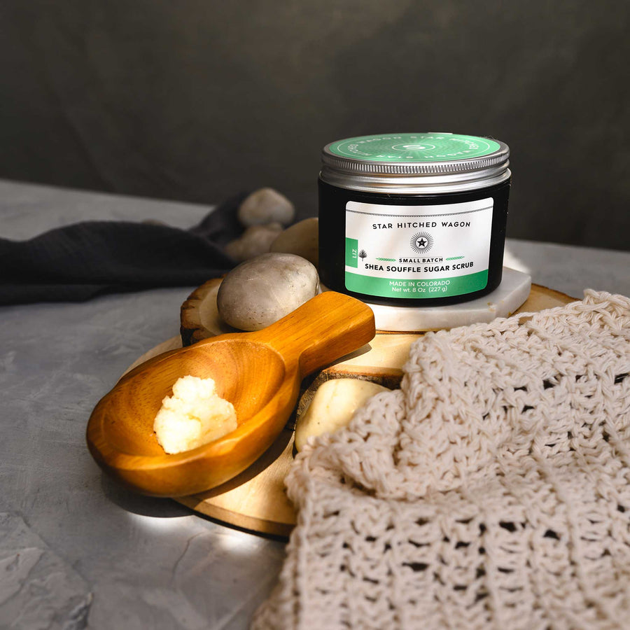A jar of Liz 8 Ounce Shea Soufflé Sugar Scrub is displayed on a wood and stone surface with a wooden spoon, knit wash cloth and small river stones.