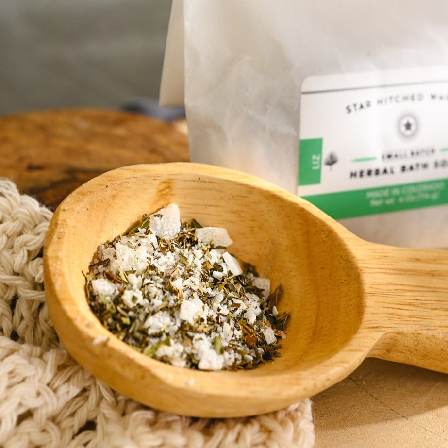 Wooden scoop filled with Liz Herbal Bath Soak next to full pouch on cream colored knit washcloth and wooden slab.