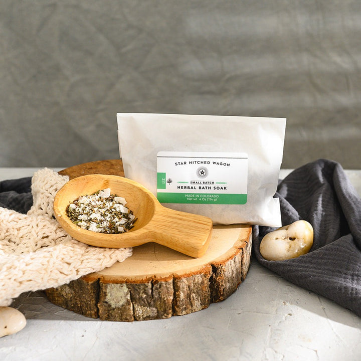 Wooden scoop filled with Liz Herbal Bath Soak next to full pouch on cream colored knit washcloth and wooden slab with grey towel and pebbles in background.