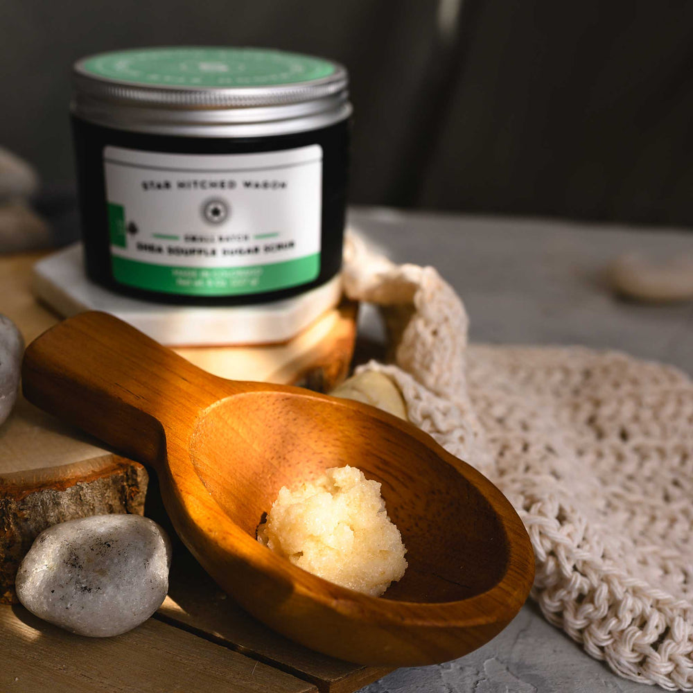 A jar of Liz 8 Ounce Shea Soufflé Sugar Scrub is displayed on a wood and stone surface with a wooden spoon, knit wash cloth and small river stones.