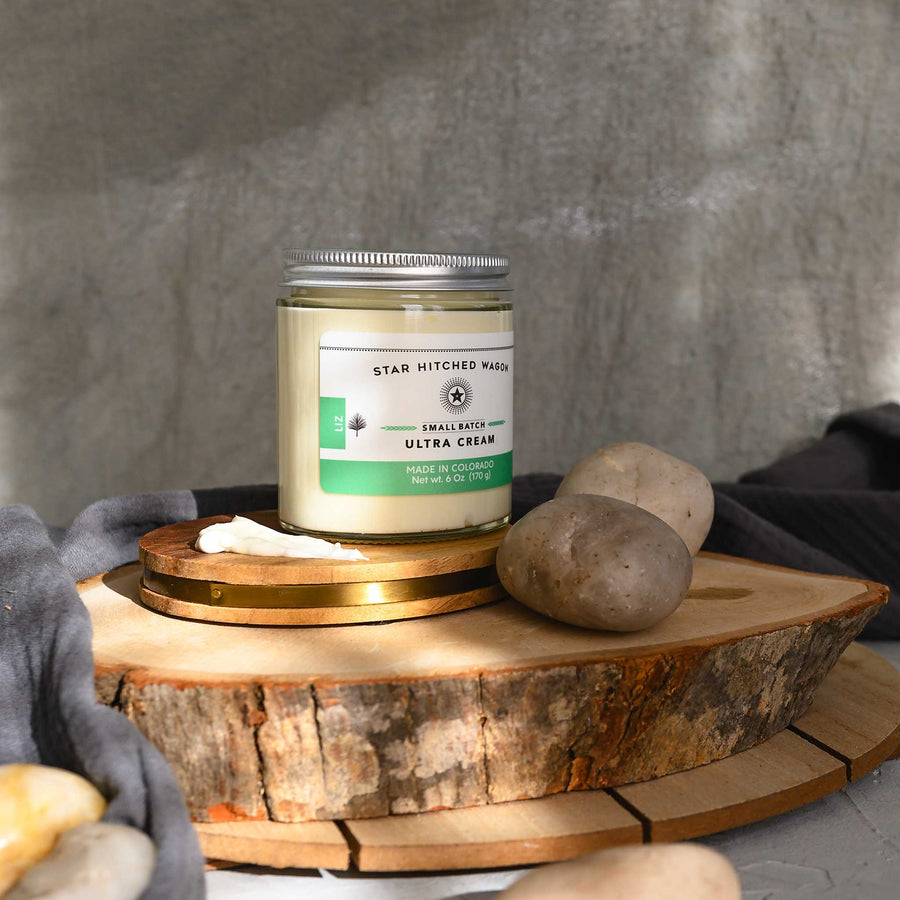 Liz Ultra Cream in a glass jar with an aluminum screw top lid, displayed on a wooden surface with a grey cloth and pebbles in the background.