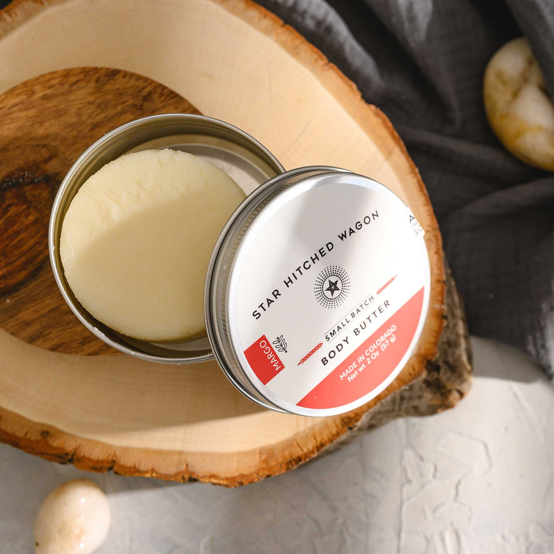 Margo body butter moisturizer in reusable tin, handcrafted with rich oils and butters to soothe dry skin, scented with citrus, herbs, and spruce, on stone and wood surface with pebbles and grey towel in background.