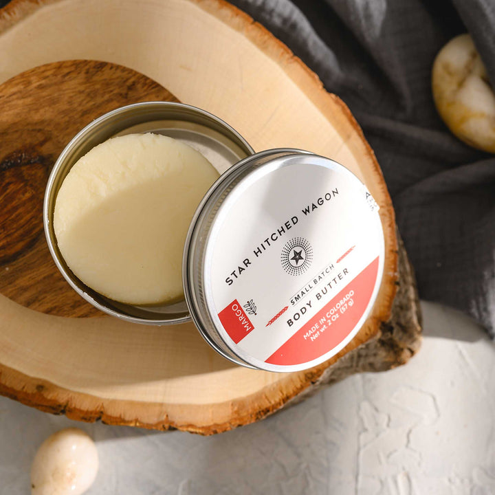 Margo body butter moisturizer in reusable tin, handcrafted with rich oils and butters to soothe dry skin, scented with citrus, herbs, and spruce, on stone and wood surface with pebbles and grey towel in background.