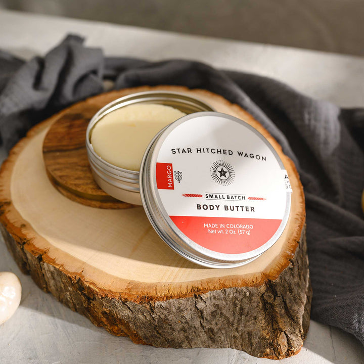 Margo body butter moisturizer in reusable tin, handcrafted with rich oils and butters to soothe dry skin, scented with citrus, herbs, and spruce, on stone and wood surface with pebbles and grey towel in background.