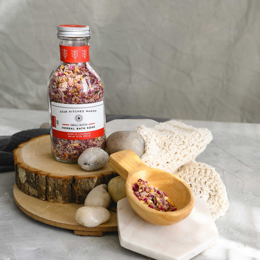 A glass bottle of Margo herbal bath soak with a screw top lid, placed on a wooden surface, next to a knit washcloth, a wooden scoop and small river stones.