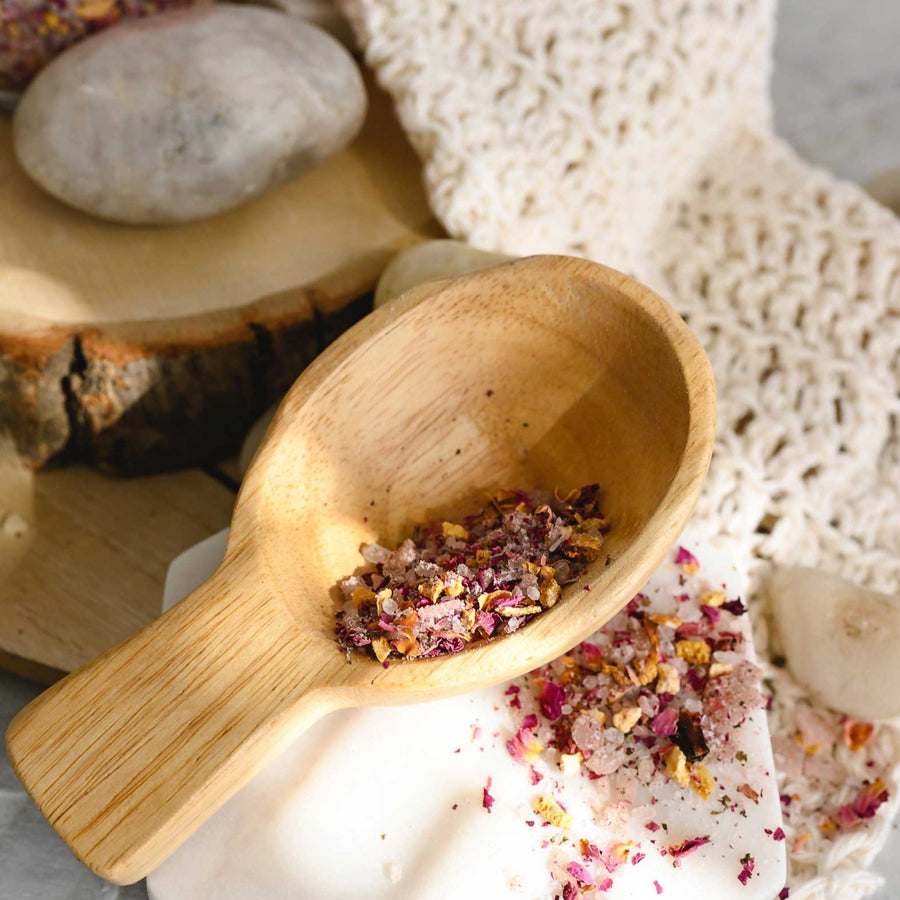 Wooden scoop filled with Margo Herbal Bath Soak on cream colored knit washcloth next to wooden slab and pebbles.