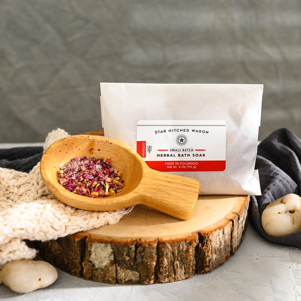 Wooden scoop filled with Margo Herbal Bath Soak next to full pouch on cream colored knit washcloth and wooden slab with grey towel and pebbles in background.