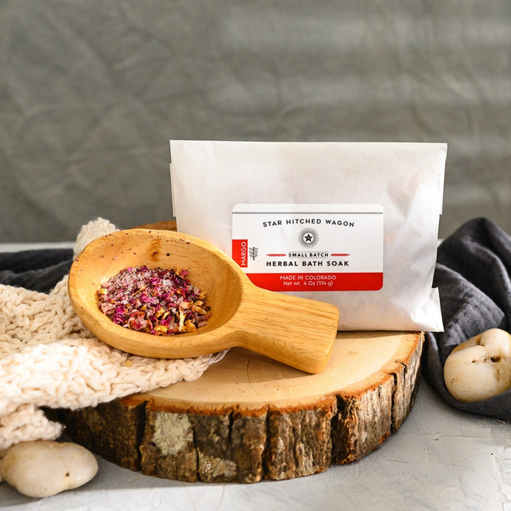Wooden scoop filled with Margo Herbal Bath Soak next to full pouch on cream colored knit washcloth and wooden slab with grey towel and pebbles in background.