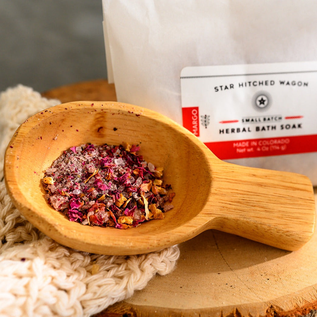 Wooden scoop filled with Margo Herbal Bath Soak next to full pouch on cream colored knit washcloth and wooden slab.