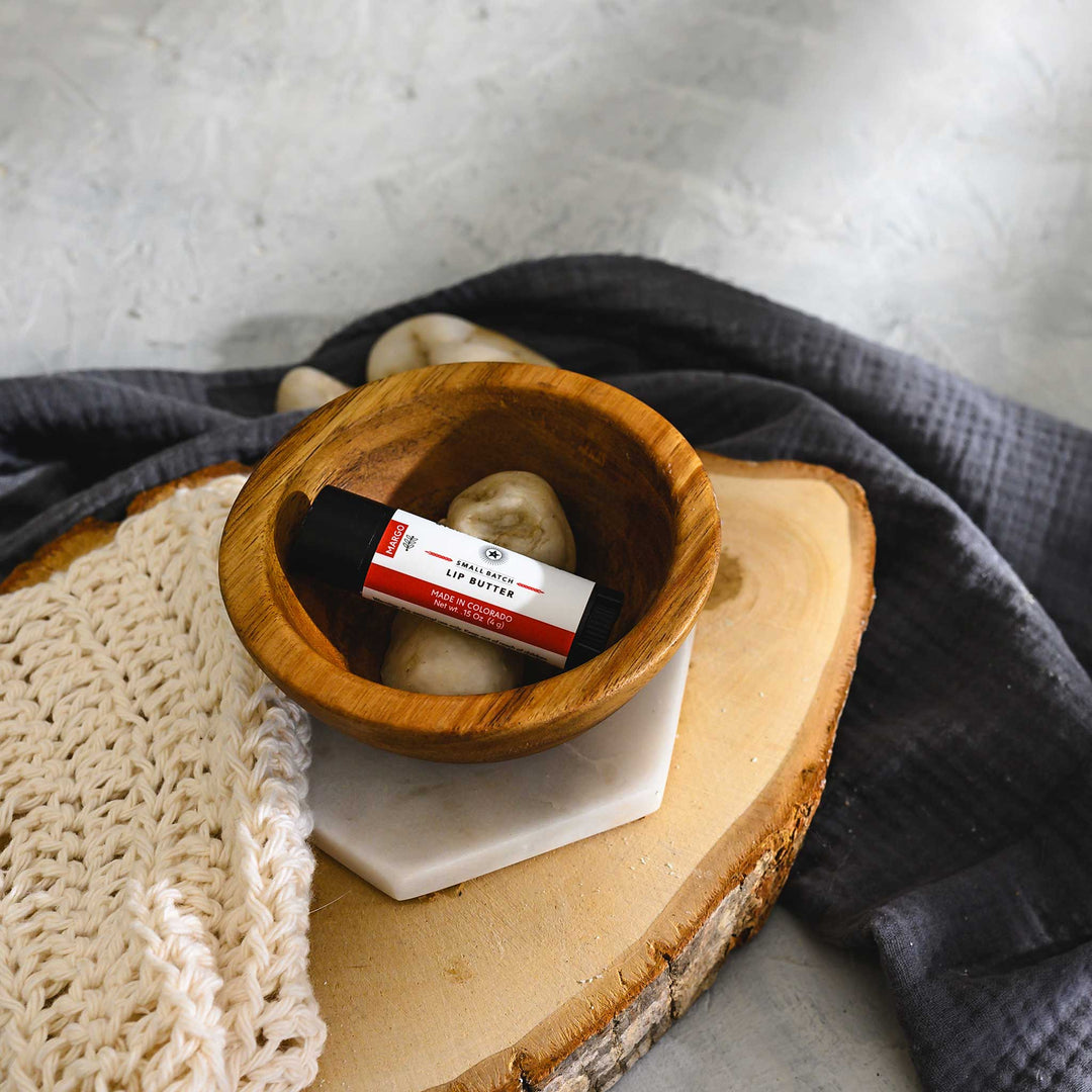A small wooden bowl with a tube of Margo Lip Butter alongside a knit washcloth on a wood and stone surface with a grey towel in the background.