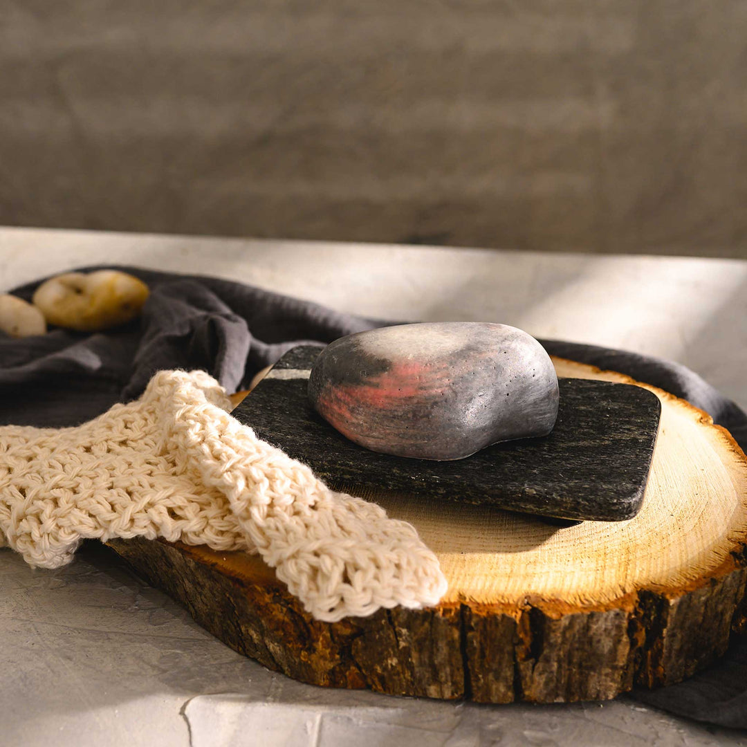 Margo river stone salt soap bar, placed on a wooden slice next to a luffa, with a dark cloth in the background.