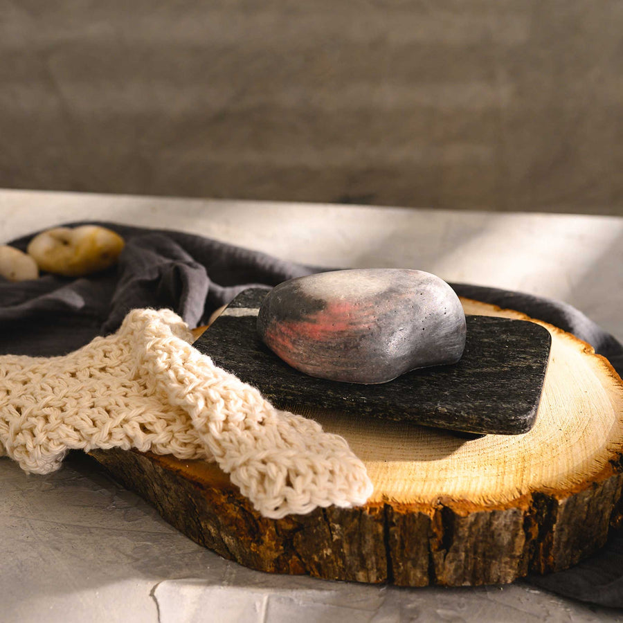 Margo river stone salt soap bar, placed on a wooden slice next to a luffa, with a dark cloth in the background.