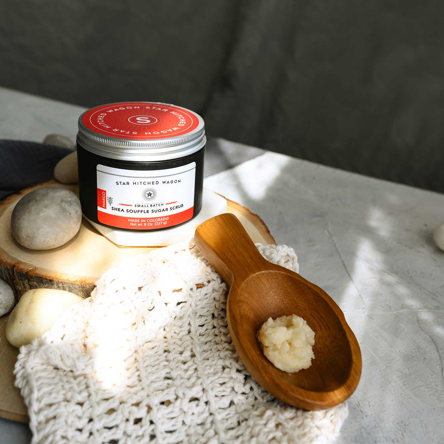 An 8 ounce jar of Margo Shea Soufflé Sugar Scrub is displayed on a wooden surface, accompanied by a pebbles, wooden scoop and a knit wash cloth.
