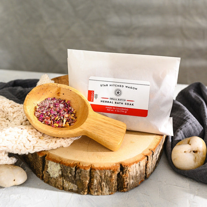 Wooden scoop filled with Margo Herbal Bath Soak next to full pouch on cream colored knit washcloth and wooden slab with grey towel and pebbles in background.