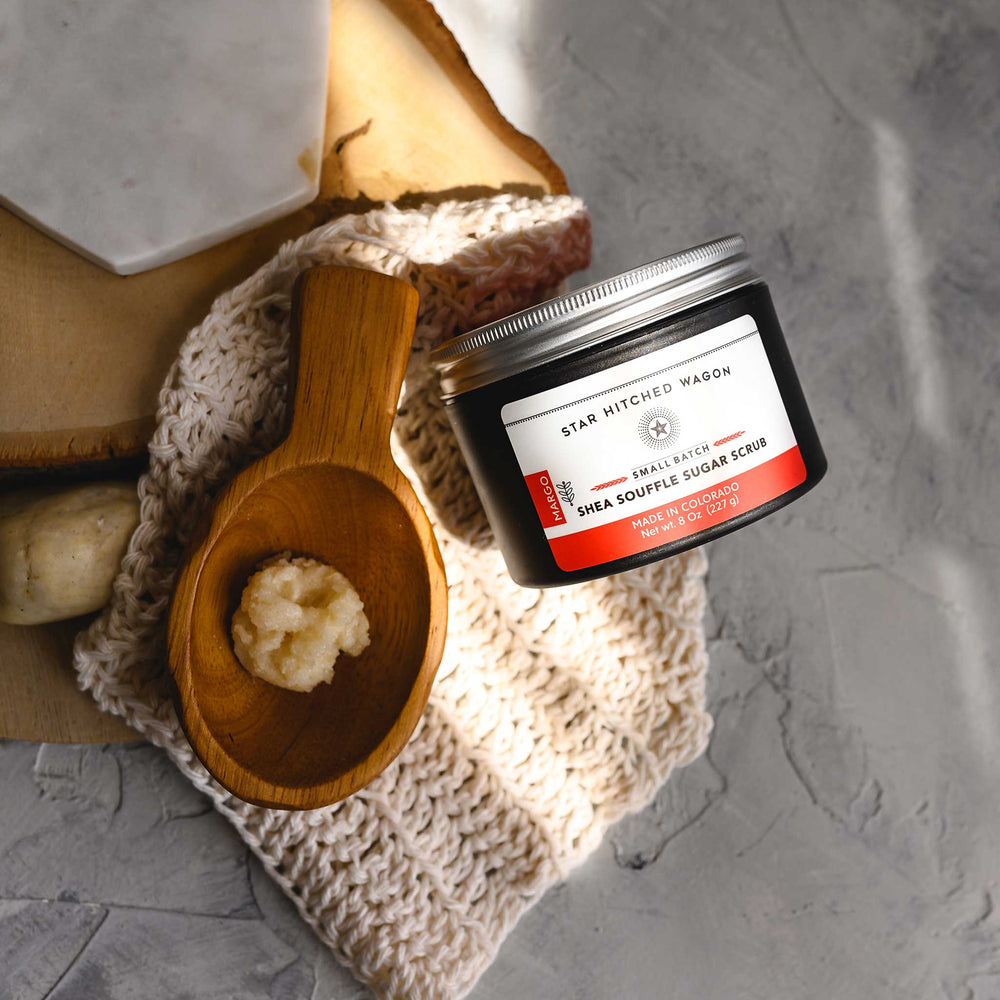 An 8 ounce jar of Margo Shea Soufflé Sugar Scrub is displayed on a wooden surface, accompanied by a pebbles, wooden scoop and a knit wash cloth.