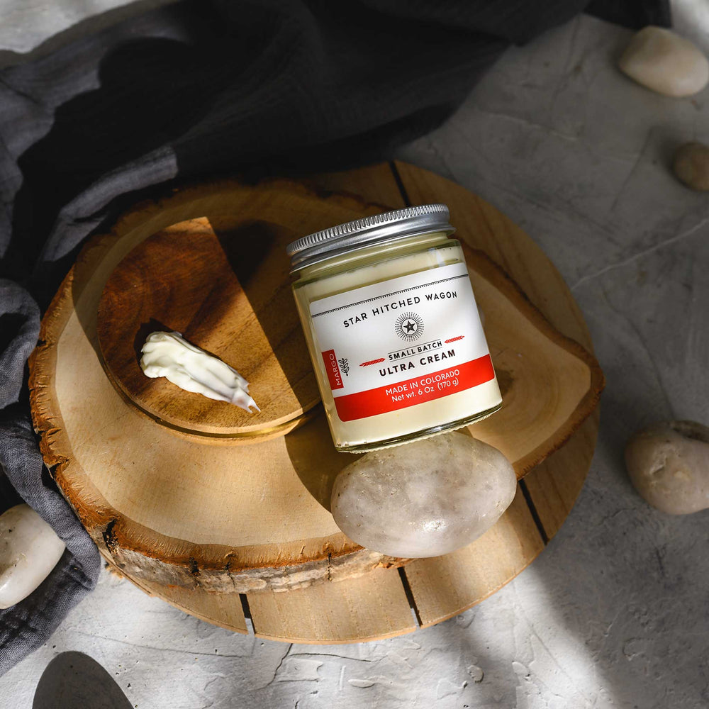 Margo Ultra Cream in a glass jar with an aluminum screw top lid, displayed on a wooden surface with a grey cloth and pebbles in the background.