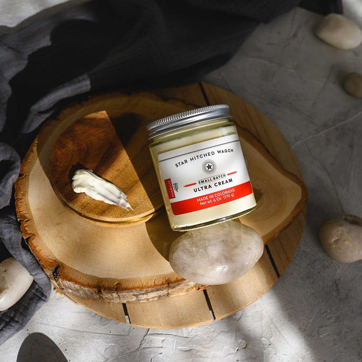 Margo Ultra Cream in a glass jar with an aluminum screw top lid, displayed on a wooden surface with a grey cloth and pebbles in the background.