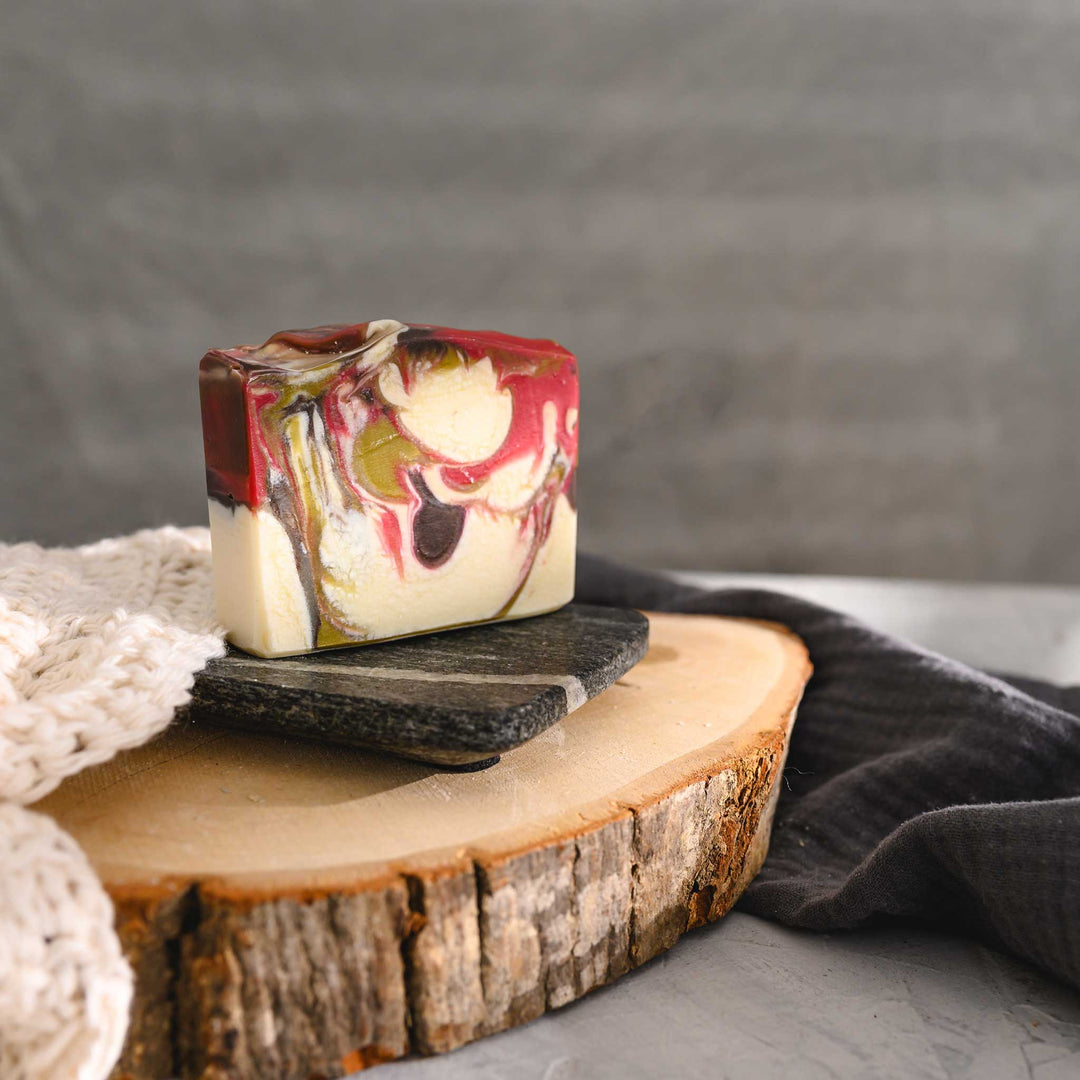 Margo essential oil soap bar with swirls of green, red, brown and white, resting on a stone and wood surface next to a cream colored washcloth with a grey towel in the background.