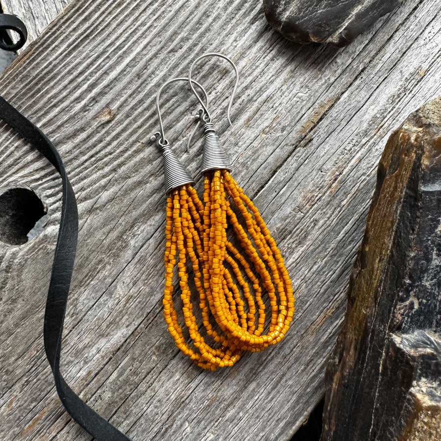 A pair of mustard yellow seed bead earrings in loop style attached to silver earwires, displayed on a wooden surface.
