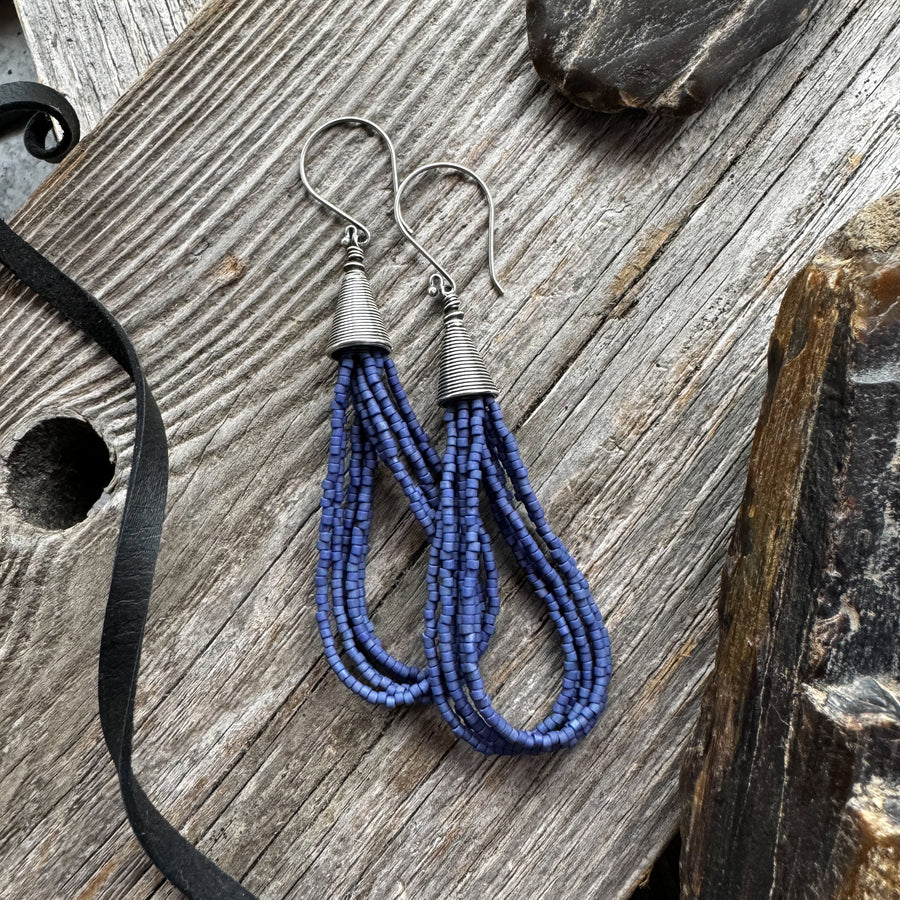 A pair of navy blue seed bead earrings in loop style with silver accents, laid on a wooden surface with a black ribbon.