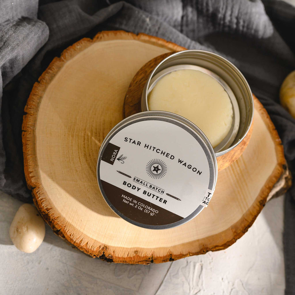 Nora body butter moisturizer in reusable tin, handcrafted with all natural oils and butters on stone and wooden surface with pebbles and grey towel in background. Sweet orange, coriander, and vanilla, like sunshine in a bottle.