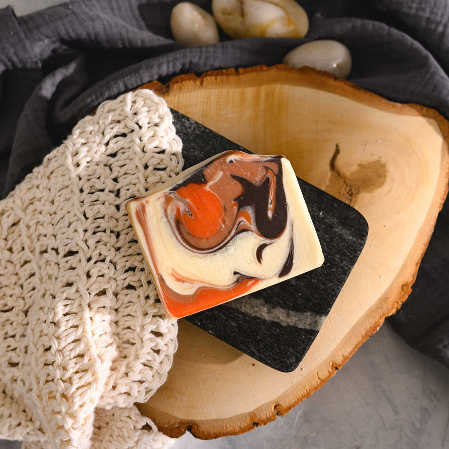 Nora essential oil soap bar with a swirl pattern in shades of orange, brown, and white, placed on a wooden surface with a crocheted washcloth beside it.