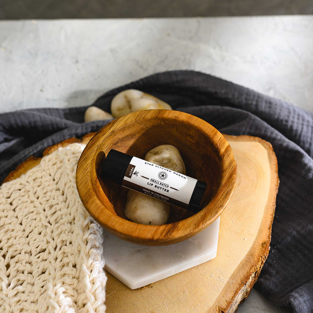 A black tube of Nora lip butter placed inside a small wooden bowl, with a crocheted washcloth, wood, stones and a grey towel in the background.