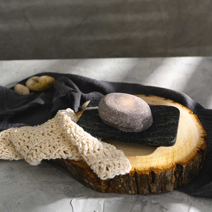 Nora river stone salt soap bar placed on a wooden slice, with a beige knitted cloth to the side.