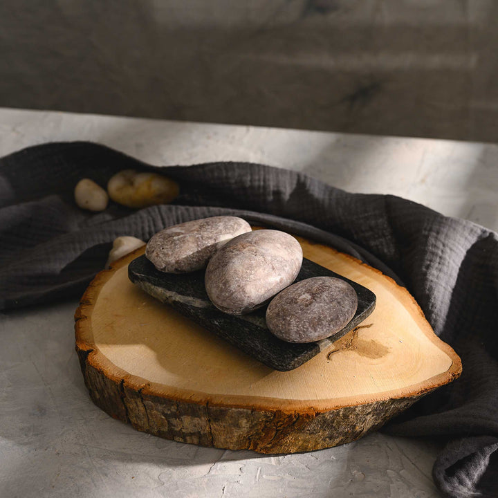 Nora river stone salt soap trio with varying shades of gray and brown, placed on a wooden disc next to a dark gray cloth.