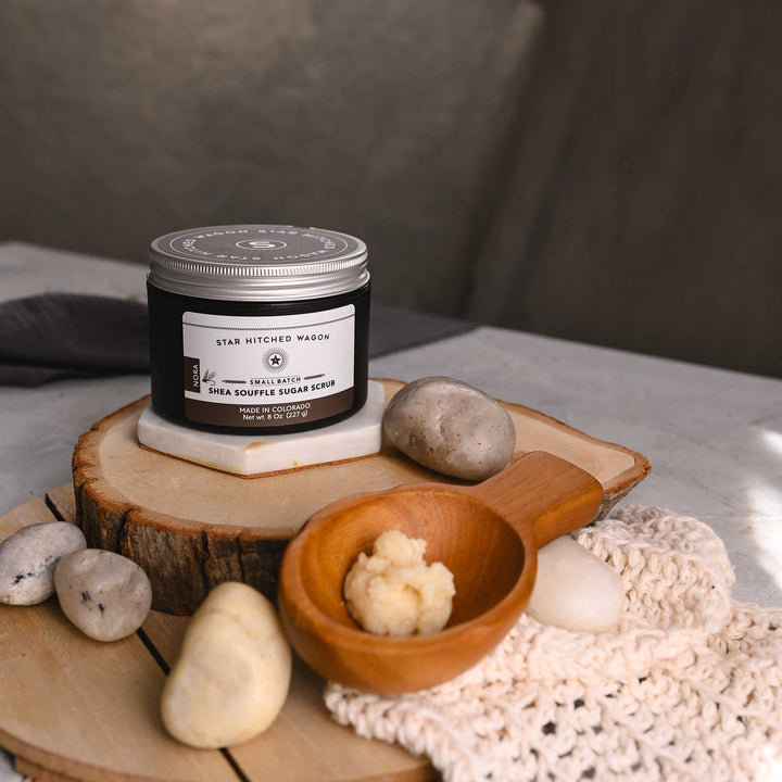 An 8 ounce jar of Nora Shea Soufflé Sugar Scrub is displayed on a wood and stone surface, accompanied by a pebbles, wooden scoop and a knit wash cloth.