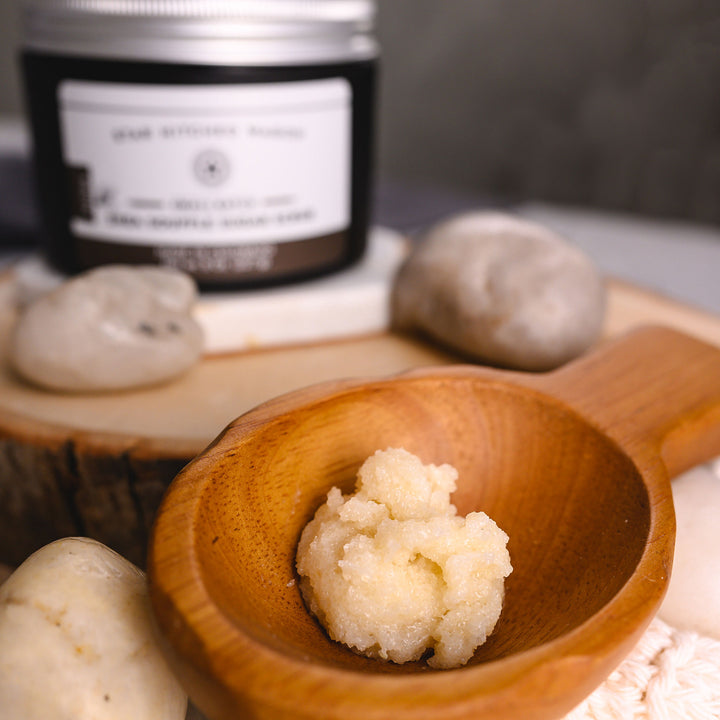 A close up of Nora Shea Soufflé Sugar Scrub is displayed in a wooden scoop with a full jar, wooden slab and pebbles in the background.