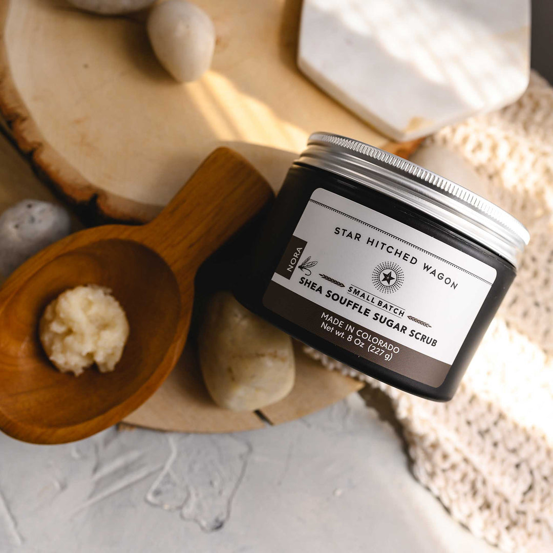 An 8 ounce jar of Nora Shea Soufflé Sugar Scrub is displayed on a wood and stone surface, accompanied by a pebbles, wooden scoop and a knit wash cloth.